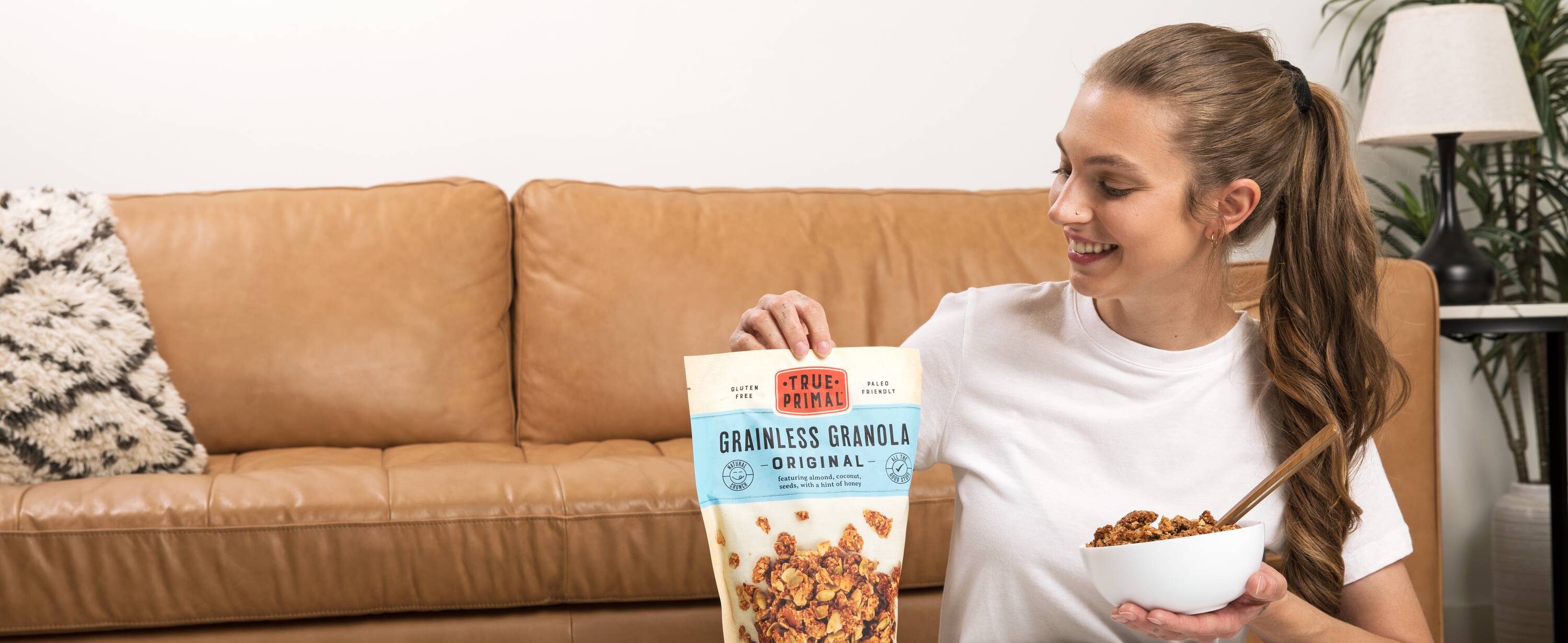 Woman eating True Primal Grainless Granola Original out of bowl next to pouch