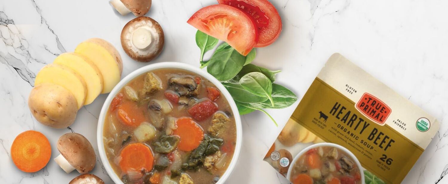 True Primal Hearty Beef Organic Soup in bowl next to vegetables and pouch