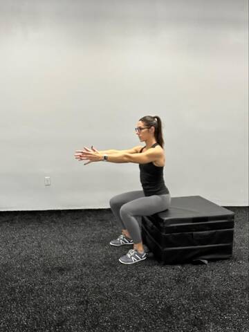 Woman in bottom position of a squat, hips resting on box for support.