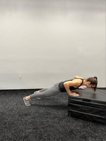 Woman in the bottom of an inclined push up. Hands under shoulders elevated to a box, straight body, elbows bending about 45 degrees away from the body.